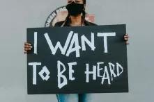Eine Frau trägt eine schwarze Maske und trägt ein Schild mit "I want to be heard" in weißer Schrift drauf.
